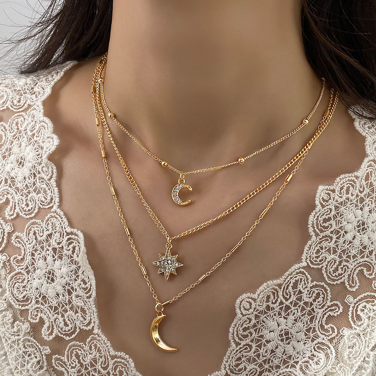 A close-up of a model wearing a layered gold necklace set with a crescent moon, star, and bead chain over a white lace top.