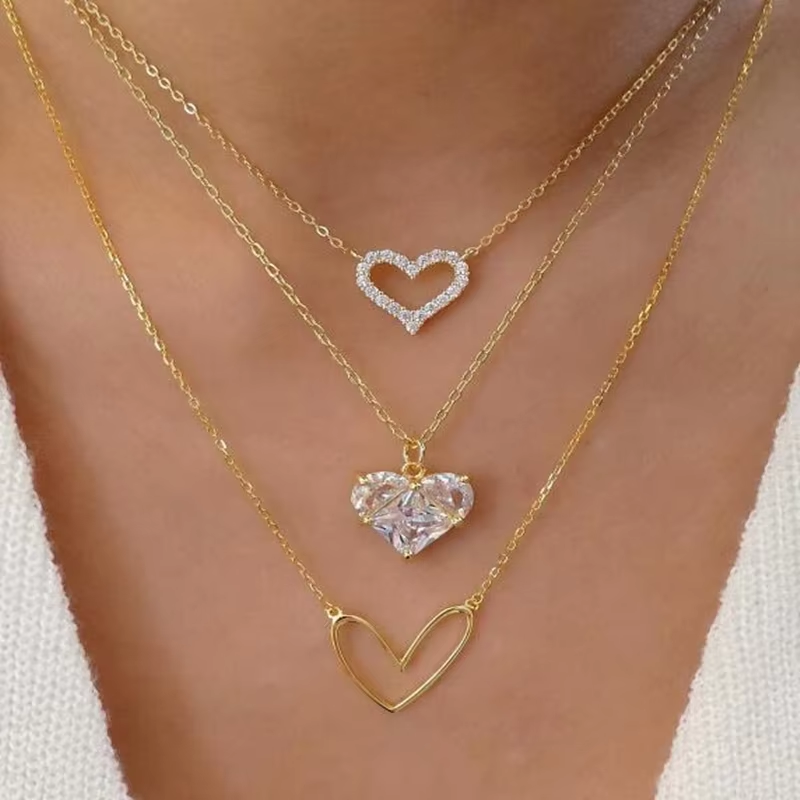 Three layered gold necklaces with heart pendants worn on a model's neck. The top necklace has a small heart outline paved with clear stones.