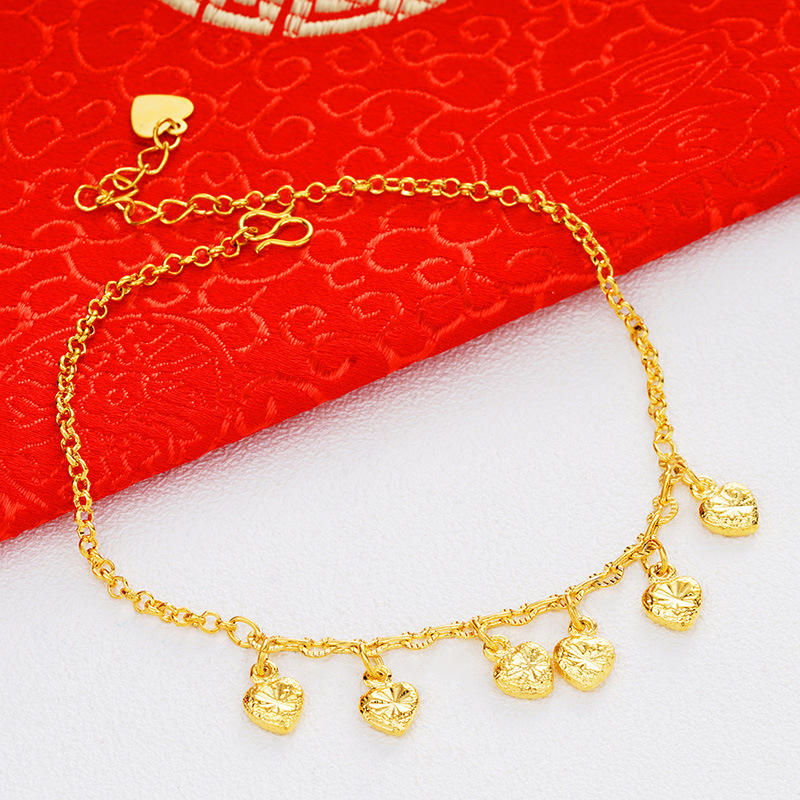 A gold chain anklet with five small textured heart charms, displayed on a white surface with a red patterned fabric background.