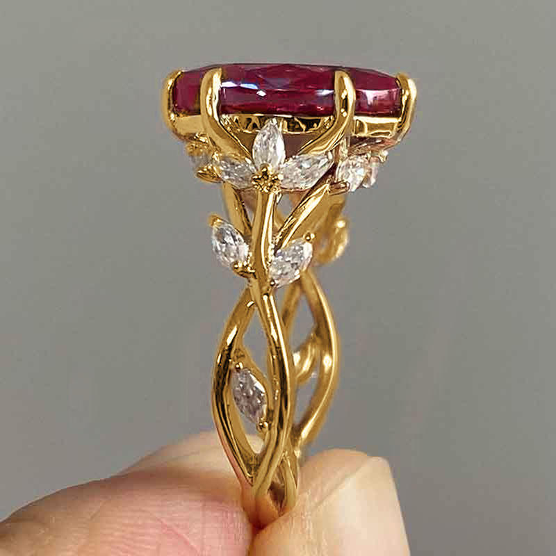 Gold ring with a red gemstone and diamond accents held by a hand against a gray background
