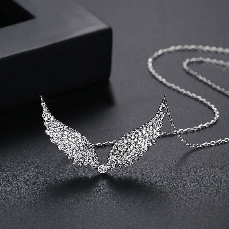 A silver-toned angel wing pendant necklace paved with clear crystals, displayed on a dark textured surface with a black box in the background.