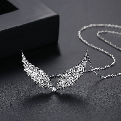 A silver-toned angel wing pendant necklace paved with clear crystals, displayed on a dark textured surface with a black box in the background.