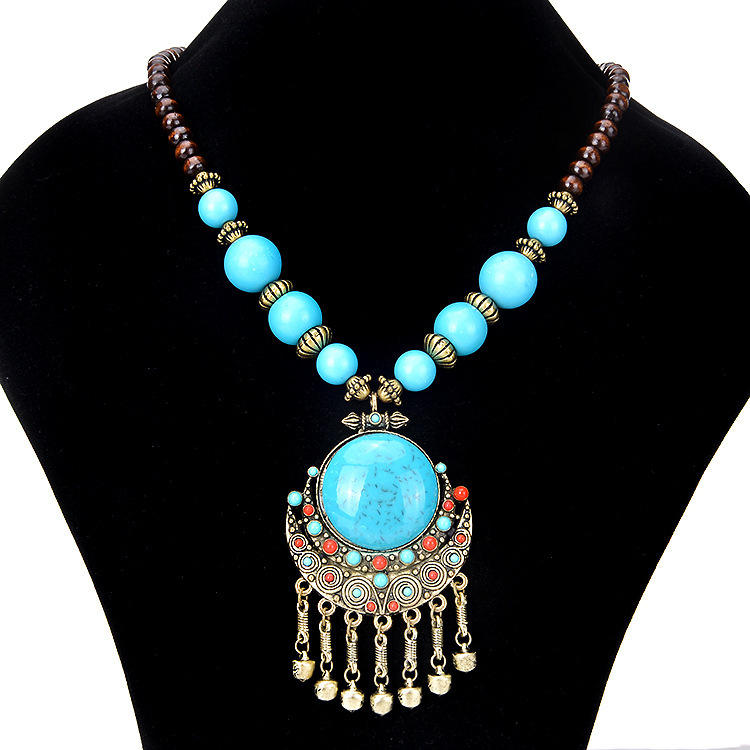 A bohemian-style statement necklace displayed on a black velvet jewelry bust. The necklace features a large central turquoise-blue stone pendant set in ornate gold-toned metal with dangling gold bells, flanked by smaller turquoise and red beads, and connected to a strand of light blue and dark brown beads with gold spacer accents.