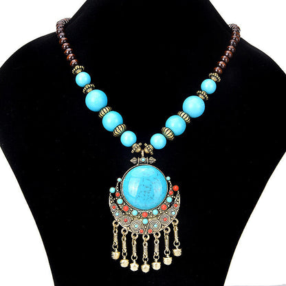 A bohemian-style statement necklace displayed on a black velvet jewelry bust. The necklace features a large central turquoise-blue stone pendant set in ornate gold-toned metal with dangling gold bells, flanked by smaller turquoise and red beads, and connected to a strand of light blue and dark brown beads with gold spacer accents.