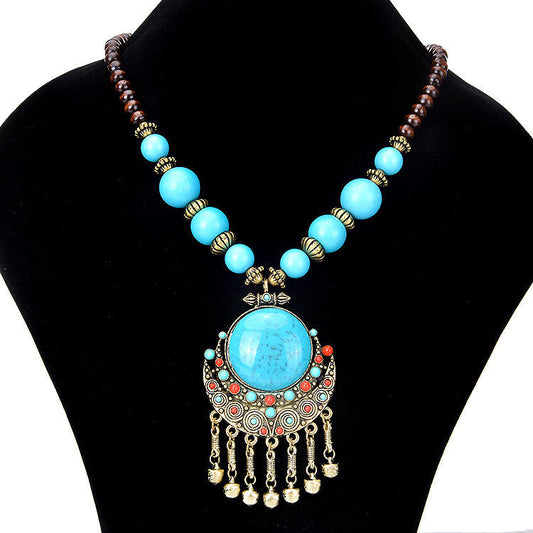 A bohemian-style statement necklace displayed on a black velvet jewelry bust. The necklace features a large central turquoise-blue stone pendant set in ornate gold-toned metal with dangling gold bells, flanked by smaller turquoise and red beads, and connected to a strand of light blue and dark brown beads with gold spacer accents.