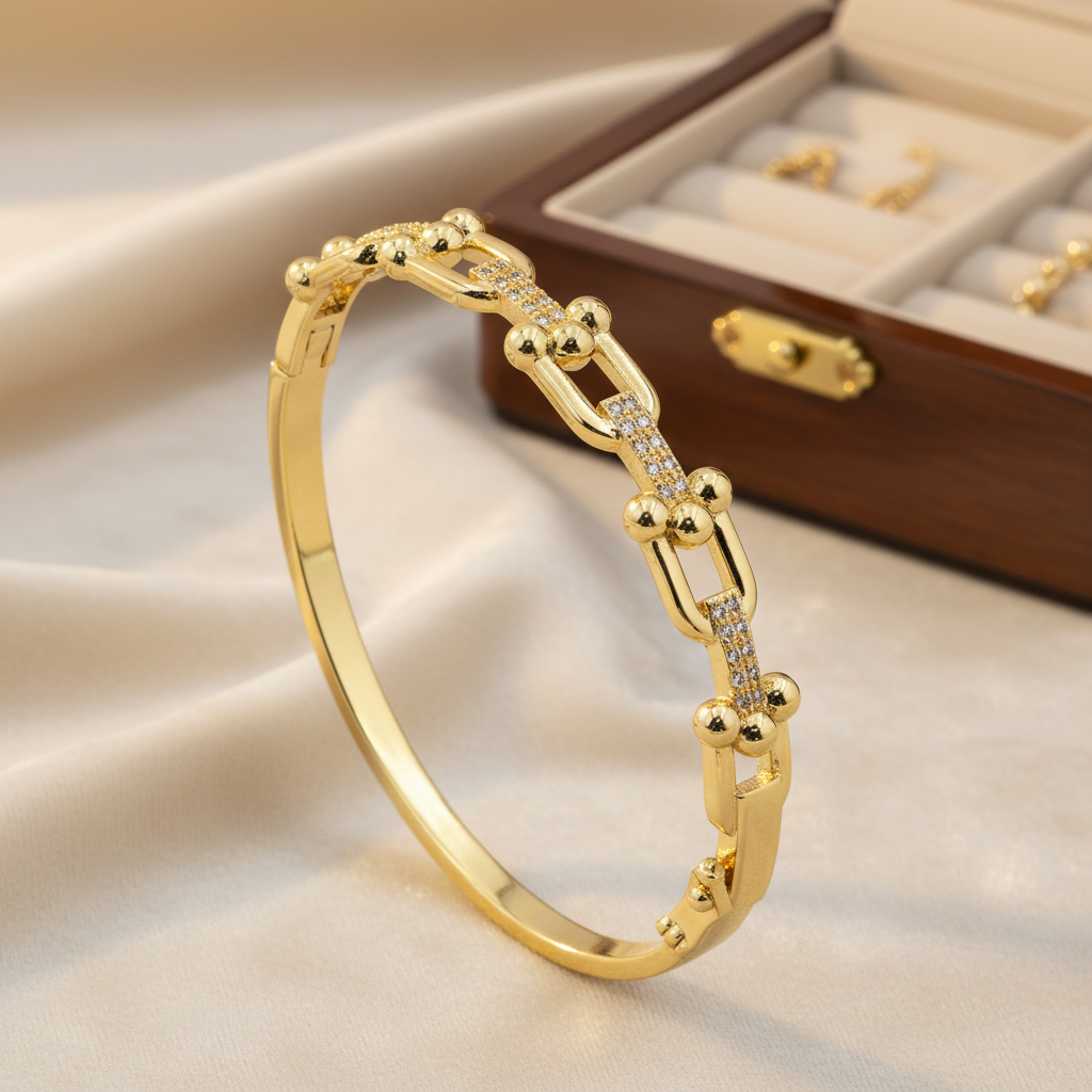 A gold hinged bangle bracelet with a chain-link design and small clear stones, displayed on a beige satin fabric next to a wooden jewelry box.