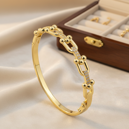 A gold hinged bangle bracelet with a chain-link design and small clear stones, displayed on a beige satin fabric next to a wooden jewelry box.