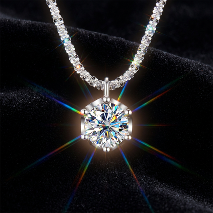 A silver-tone necklace with a large round brilliant-cut clear gemstone pendant, displayed on a black velvet background with rainbow light flares.