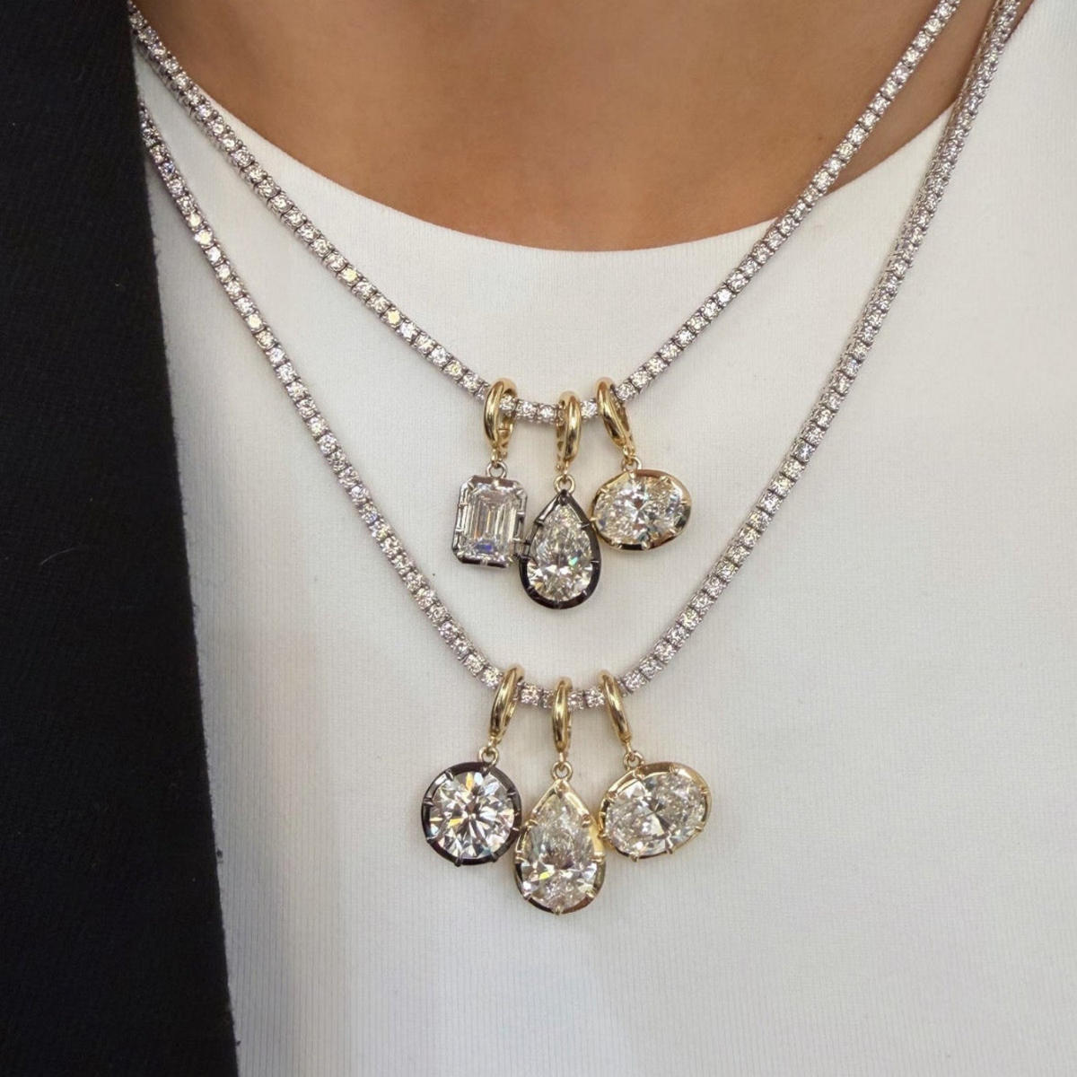 A close-up of a person wearing a layered necklace set. The set includes a silver chain with a cluster of clear baguette and oval stones, and a gold chain with a cluster of clear teardrop and oval stones, both resting on a white top.
