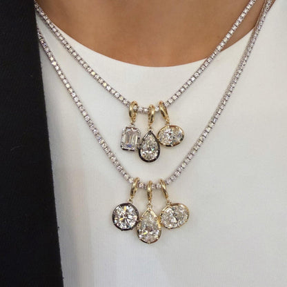 A close-up of a person wearing a layered necklace set. The set includes a silver chain with a cluster of clear baguette and oval stones, and a gold chain with a cluster of clear teardrop and oval stones, both resting on a white top.