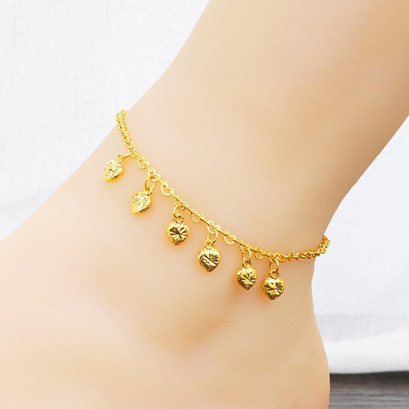 A delicate gold anklet with five small textured heart charms, shown worn on a person's ankle against a soft white background.