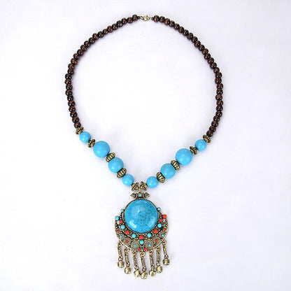A statement necklace featuring a large oval turquoise-blue stone pendant with dangling gold-toned fringe, accented by small turquoise and red beads, and a dark brown beaded chain on a white background.