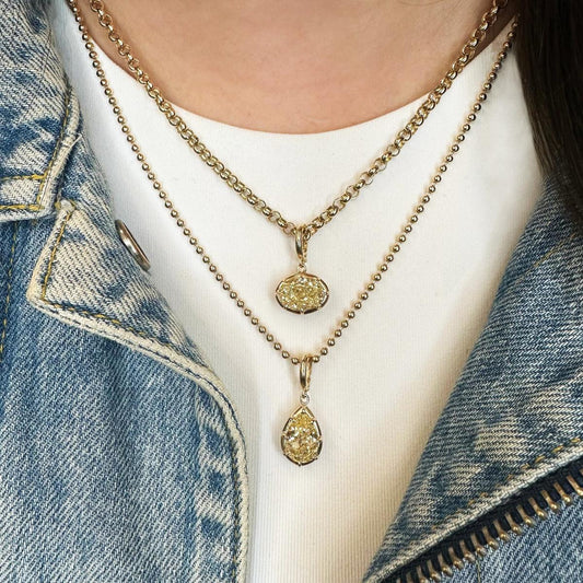 A close-up of a person wearing layered gold necklaces, including a curb chain with an oval textured pendant and a beaded chain with a teardrop pendant, over a white top and blue denim jacket.