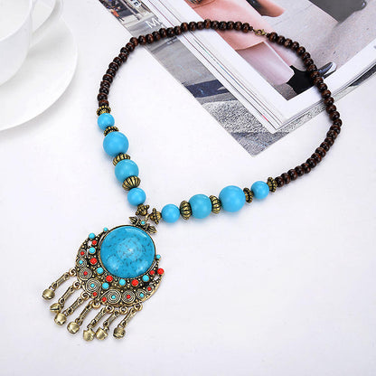 A bohemian-style statement necklace featuring a large turquoise-blue stone pendant with dangling gold-toned tassels, accented by turquoise beads, small red beads, and dark brown beads on a white background with a magazine and teacup prop.