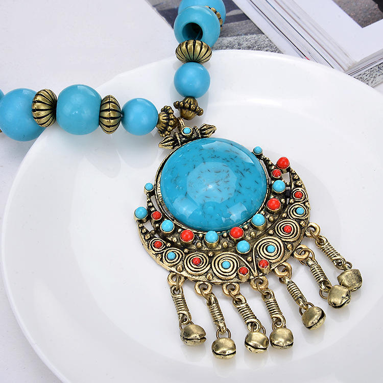 A close-up of a bohemian-style turquoise and gold statement necklace resting on a white plate. The necklace features a large round turquoise cabochon pendant surrounded by intricate gold filigree, small red and turquoise accent stones, and five dangling gold bell charms.