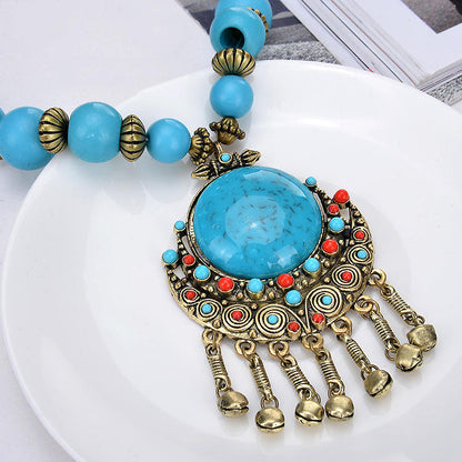 A close-up of a bohemian-style turquoise and gold statement necklace resting on a white plate. The necklace features a large round turquoise cabochon pendant surrounded by intricate gold filigree, small red and turquoise accent stones, and five dangling gold bell charms.
