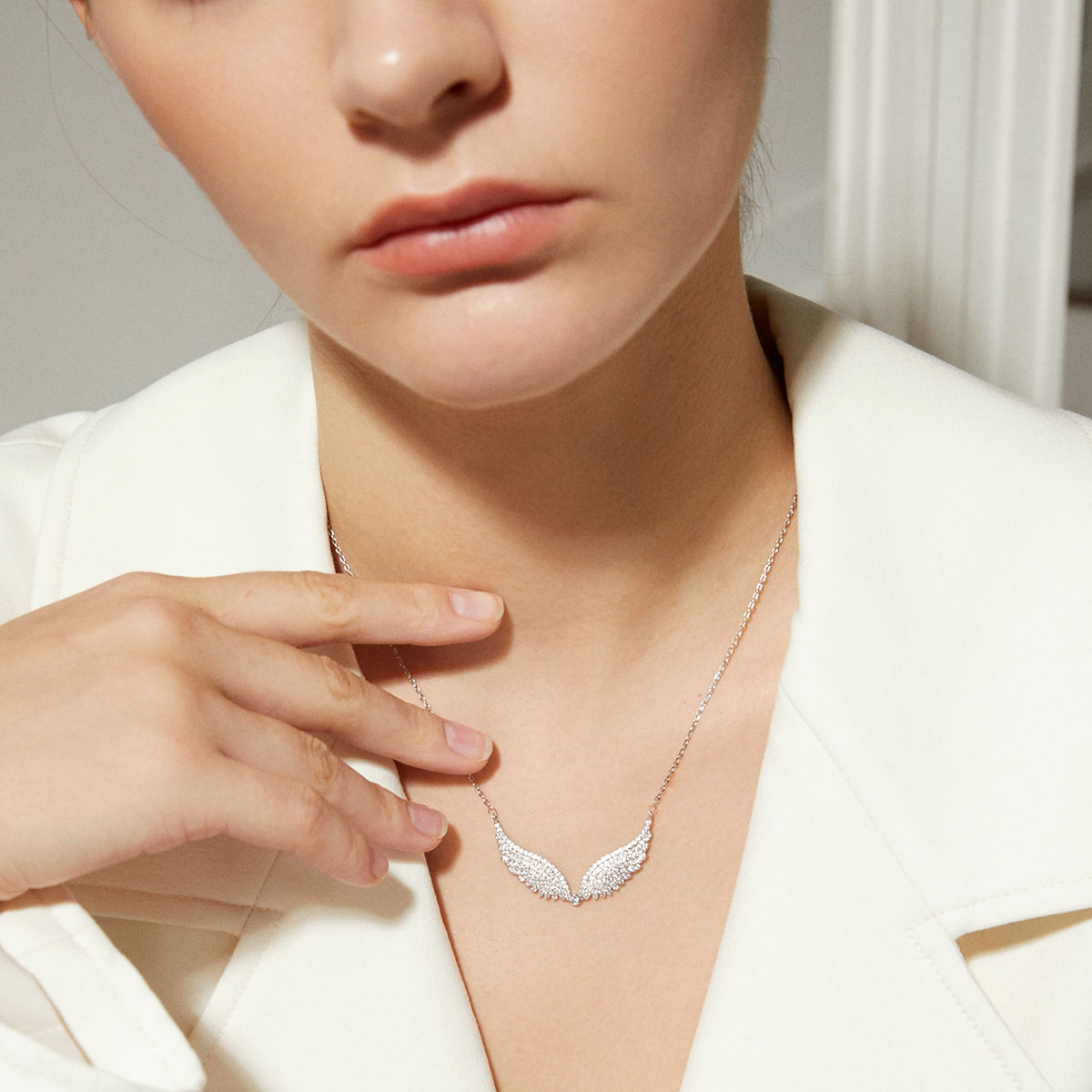 A close-up lifestyle shot of a model wearing a delicate silver wing pendant necklace. The necklace features a pair of curved, textured wings set with small clear stones, hanging from a fine silver chain.