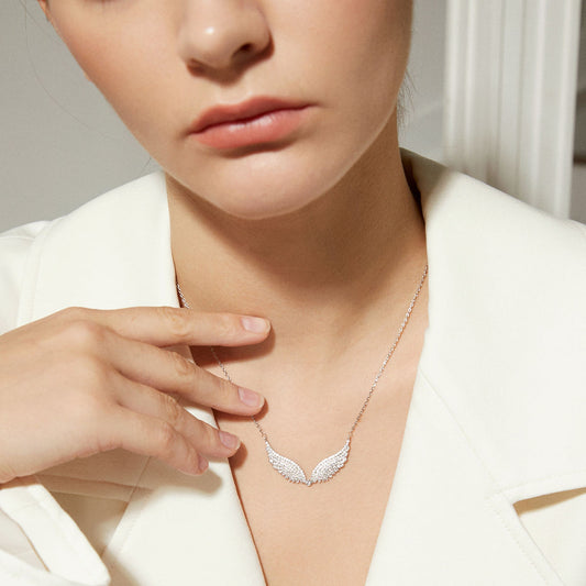 A close-up lifestyle shot of a model wearing a delicate silver wing pendant necklace. The necklace features a pair of curved, textured wings set with small clear stones, hanging from a fine silver chain.