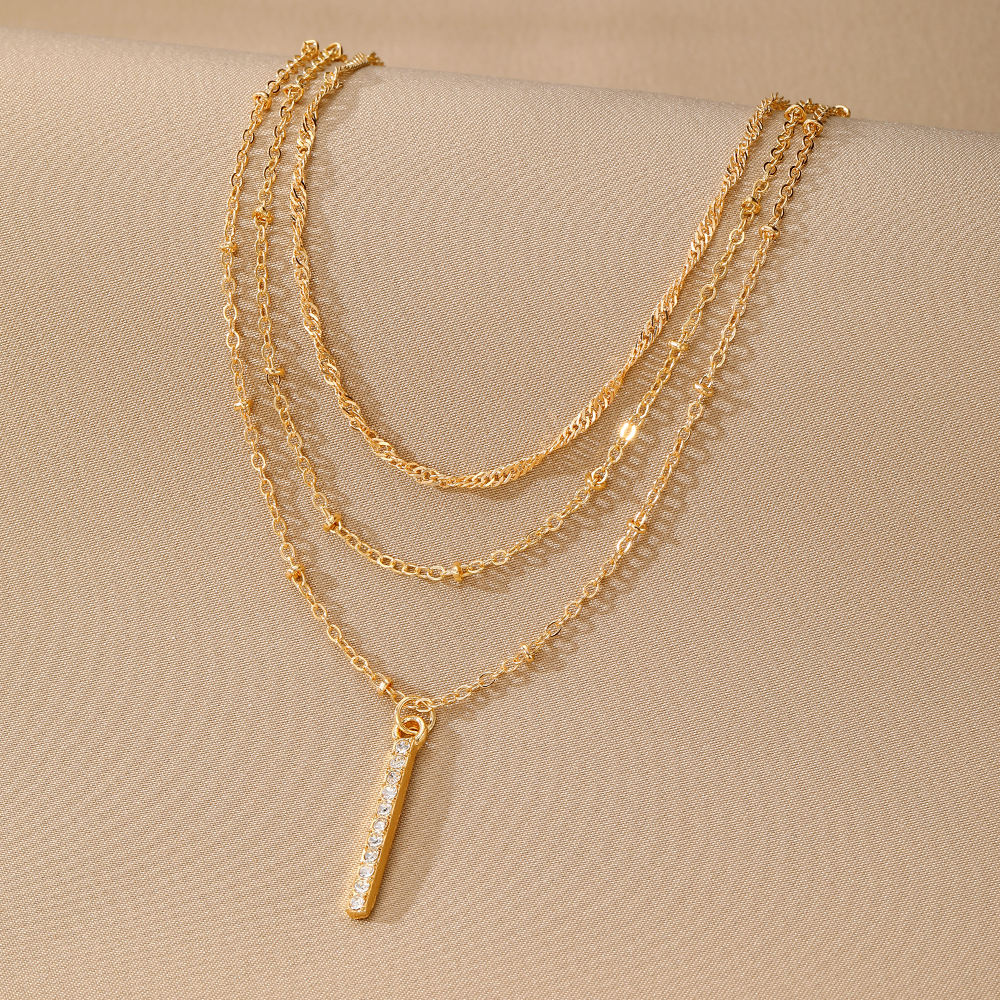 A gold multi-layer necklace with a vertical bar pendant adorned with small clear stones, displayed on a textured beige surface.