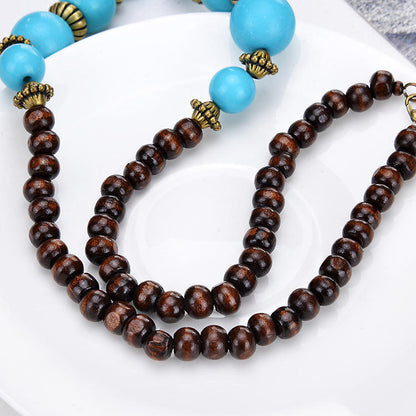 A close-up of a beaded necklace draped on a white plate. The necklace features dark brown wooden beads with antique bronze-colored metal spacers, alongside bright turquoise blue round beads.