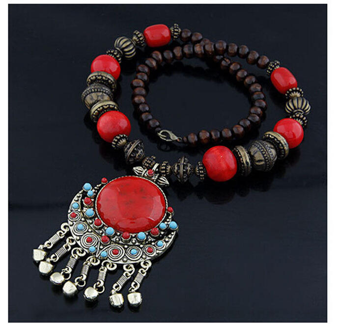 A beaded bracelet with a large ornate pendant, featuring bright red round beads, dark brown wooden beads, and silver-toned metal spacers, with a large red stone pendant and dangling silver charms on a black background.