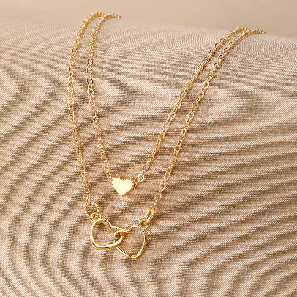 A gold layered necklace set displayed on a beige fabric surface, featuring a delicate chain with a small heart pendant and a longer chain with two interlocking open heart pendants.